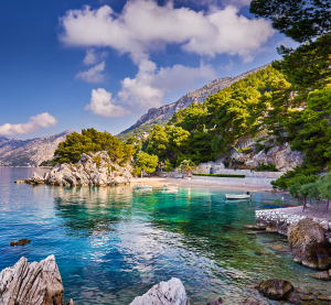 Felsige K&uuml;ste mit t&uuml;rkisfarbenem Wasser und Pinienb&auml;umen in Kroatien, im Hintergrund Berge und ein kleiner Strand mit Booten.