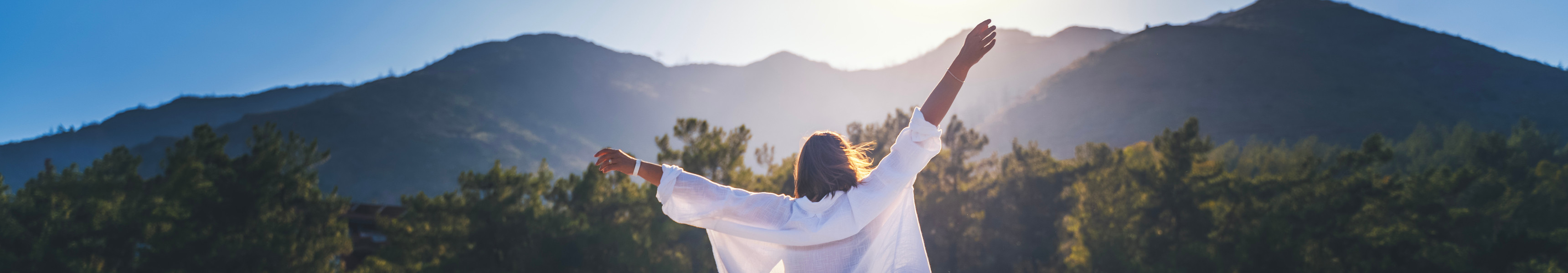 Eine Frau in einem wei&szlig;en Hemd genie&szlig;t mit erhobenen H&auml;nden die Morgensonne vor der Kulisse der Berge bei Sonnenaufgang