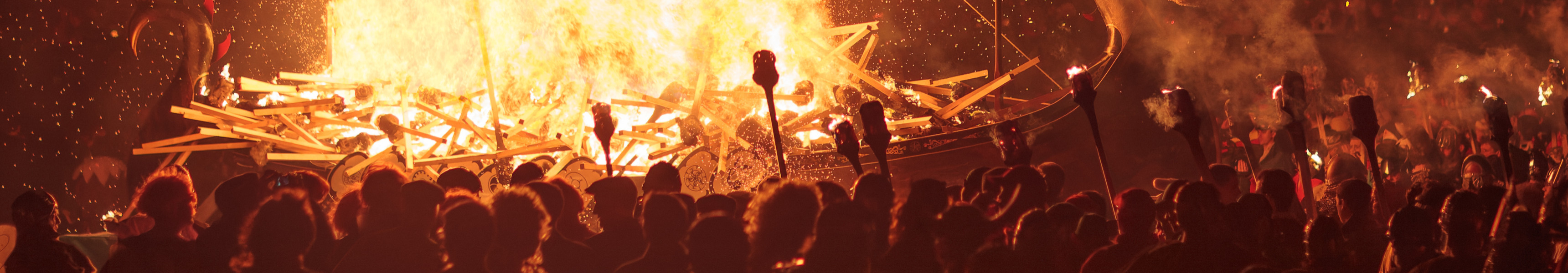 Ein gro&szlig;es, brennendes Wikingerschiff bei einem Feuerfest in der Nacht, mit vielen Zuschauern und Fackeln.