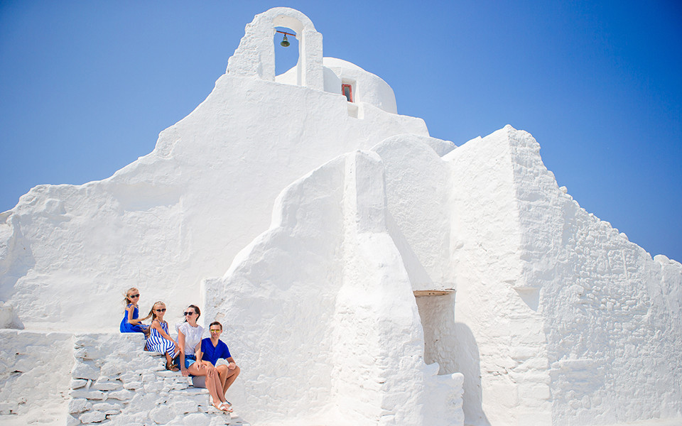Familie sitzt auf den Treppen neben einer ikonischen wei&szlig;en Kirche auf Mykonos, umgeben von strahlend blauem HImmel. 