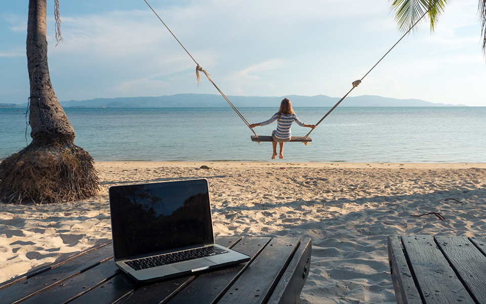 Laptop am Strand, Frau schaukelt mit Blick aufs Meer beim Digital Detox Urlaub.