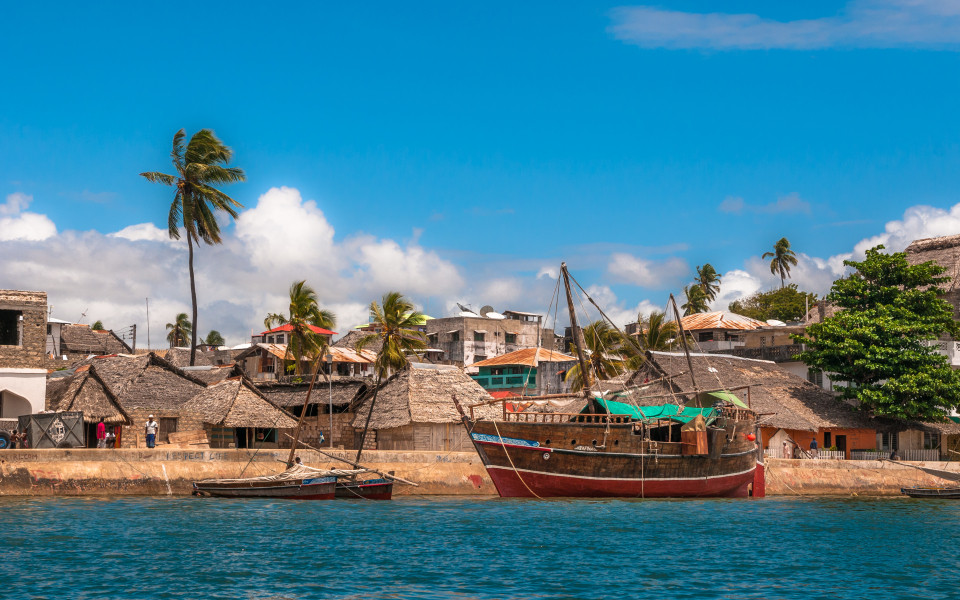 K&uuml;ste von Lamu mit traditionellen H&auml;usern und einem Holzboot im Vordergrund.